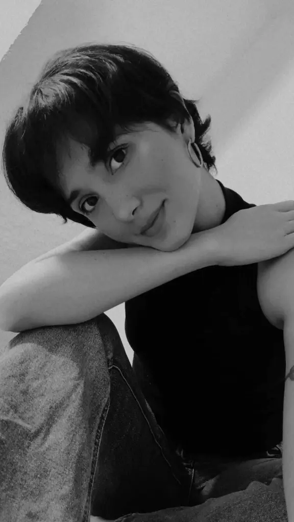 Cherrylb with short dark hair and large hoop earrings smiles while laying down in a white shirt.