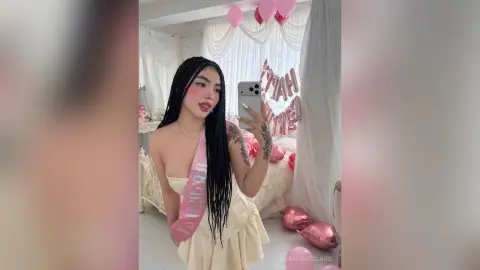 ValeriaEclaire poses in a light yellow dress with long black hair, standing in a white bedroom decorated with pink balloons.