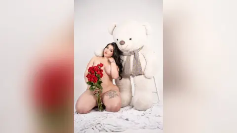 Nude woman holding red roses sitting next to a giant white teddy bear in a romantic studio setting.