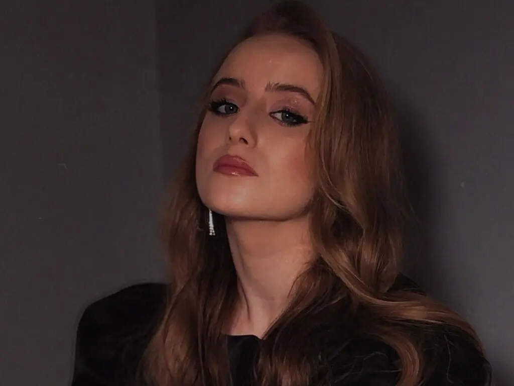 Young woman with long wavy hair wearing a black outfit posing with a moody expression against a dark background.