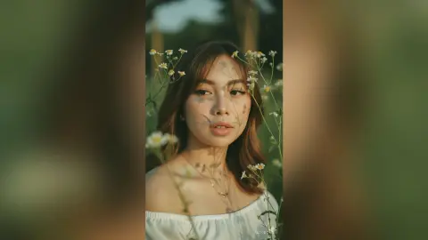 Dreamy portrait of a young woman in a white off-the-shoulder top amidst wildflowers in a sunlit meadow.