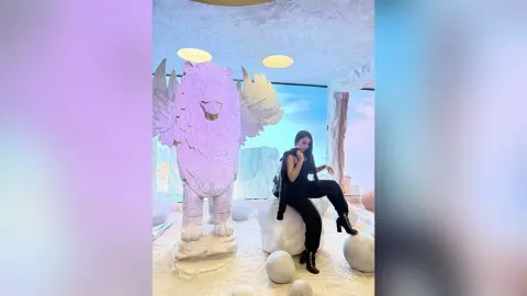 Woman in black outfit sitting by a large white winged lion statue in a dreamy, ethereal indoor setting.