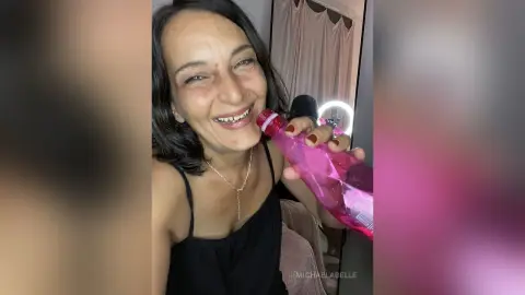 MichaelaBelle smiles while holding a pink bottle near her face, wearing a black tank top and a silver necklace.