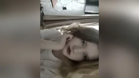 GemmaCiliberto lying on a bed with her hand resting against her cheek, looking directly at the camera with long blonde hair.