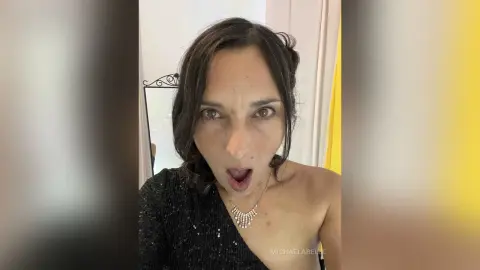 MichaelaBelle looks into the camera with an open mouth, wearing a black sparkly top and a delicate silver necklace.