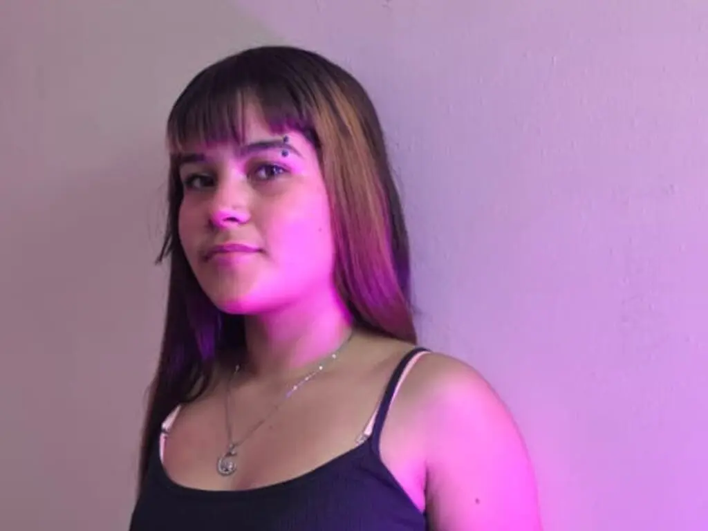 Young woman with brown hair and silver necklace wearing a black tank top in soft pink lighting.