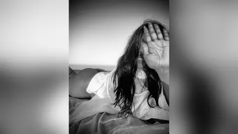 Melancholic black and white portrait of a woman in a white shirt covering her face by the seaside.
