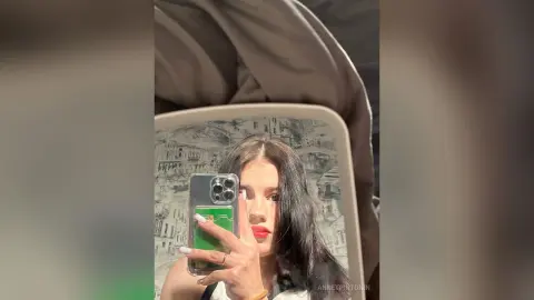 Young woman with long dark hair taking a mirror selfie in a cozy bedroom setting.