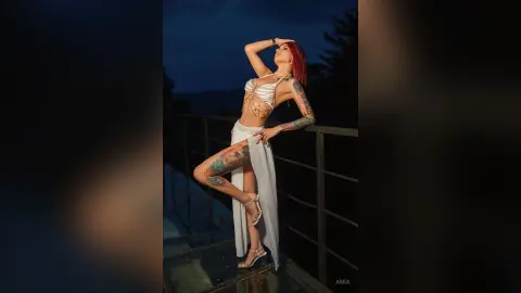Red-haired woman in a white crop top and slit skirt posing on a balcony at twilight.