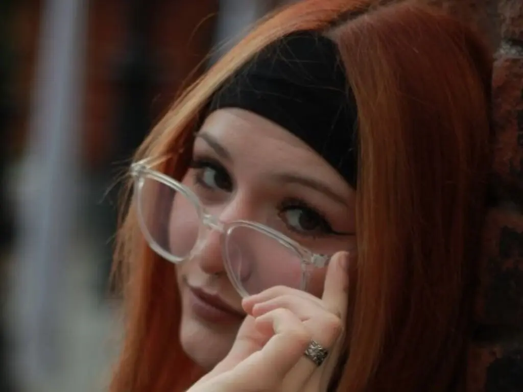 Redheaded woman with clear glasses and a black headband looking thoughtful in a soft outdoor setting.