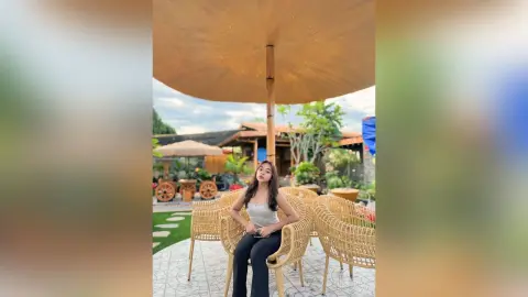 Young woman in a white tank top sitting at a wicker cafe table under a large sun umbrella on a sunny patio.