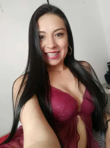 Smiling young woman with long dark hair wearing a burgundy lace dress in a bright indoor setting.