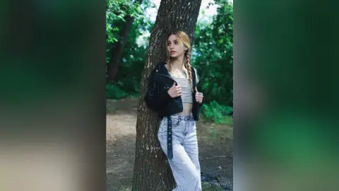 Young woman in a black leather jacket and light denim jeans leaning against a tree in a lush green forest.