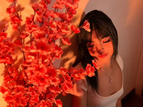 DapneOwen poses with long dark hair and heavy lashes next to bright pink flowers in warm lighting.