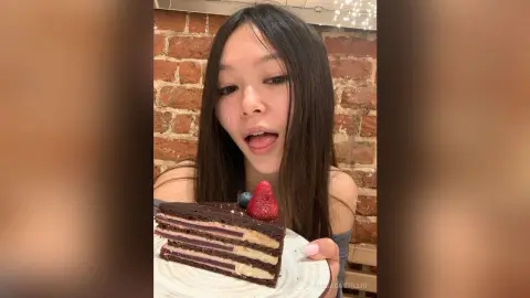 Young woman in a casual top enjoying a slice of chocolate layer cake in a rustic brick room.