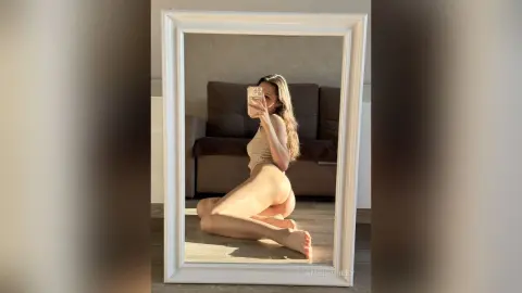 AmeliaRiley poses naked in front of a mirror, showing her bare buttocks and side profile while kneeling on the floor.