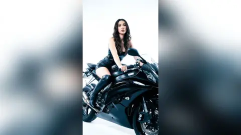 Stylish woman in black leather outfit sitting on a black sport motorcycle against a white studio background.