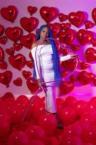 Woman in white dress holding heart wand amidst red heart balloons in a romantic pink studio setting.