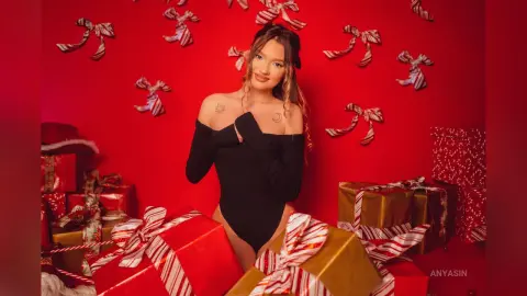 Smiling woman in a black bodysuit posing with red and gold Christmas gifts against a festive red background.