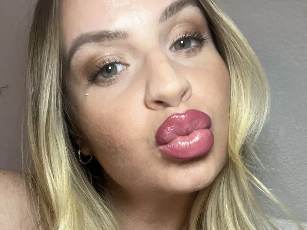 Blonde woman with glamorous makeup and pink lipstick making a playful pouty face in a close-up portrait.