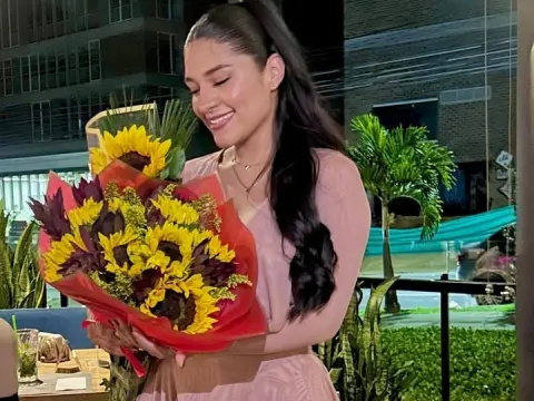 Smiling woman in a pink dress holding a large bouquet of sunflowers on a rooftop balcony at night.