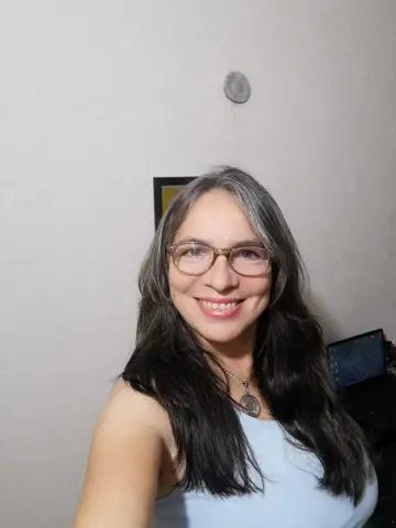 Smiling woman with long grey hair wearing glasses and a light blue tank top in a bright indoor setting.