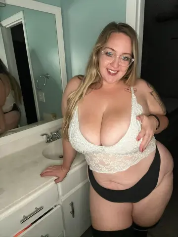 Smiling woman in white lace lingerie and black panties posing in a bright bathroom.