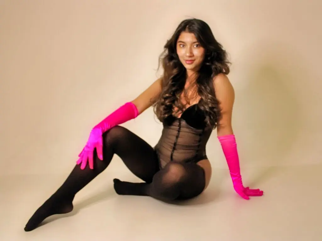 MadiisonWilliams wears a black sheer bodysuit and long pink gloves while posing on the floor against a plain background.
