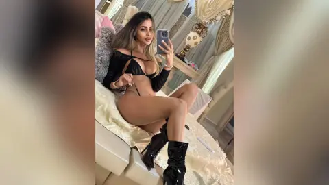 MiaGuerra poses on a bed wearing a black bra and thong, showing off her long hair and black boots.