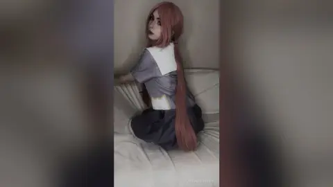 Cosplayer with long pink hair wearing a grey and white anime school uniform sitting on a white bed.
