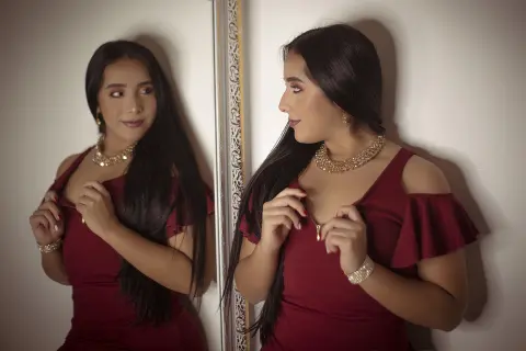Elegant woman in a burgundy cocktail dress and gold necklace posing in front of a mirror.