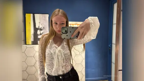 AdenaMcfalls takes a mirror selfie wearing a white lace long-sleeve blouse and black pants, showing her long blonde hair and light complexion.