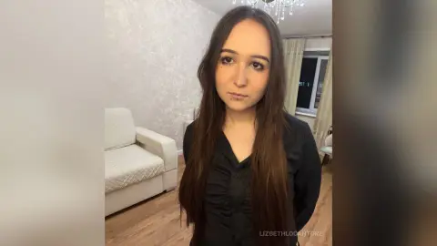 LizbethLocantore looks at the camera with long brown hair, wearing a black button-up shirt in a room with a white sofa and light walls.