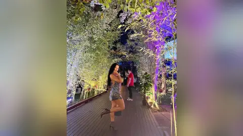 NicoleBella stands on a wooden walkway wearing a short, patterned silver dress and black high heels, surrounded by lush green and purple foliage.