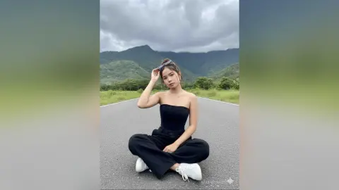 Young woman in a black strapless top and trousers sitting on a road against a scenic mountain landscape.