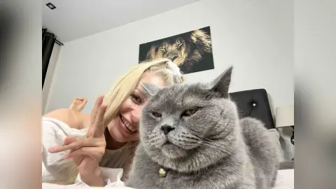 Smiling blonde woman in a white top cuddling a large grey British Shorthair cat in a cozy bedroom setting.