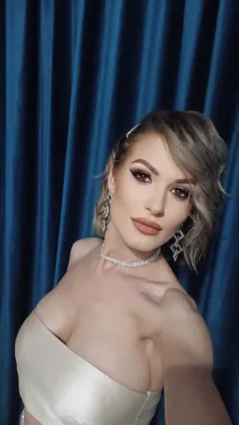 Glamorous woman in a white strapless dress and diamond necklace posing against blue velvet curtains.