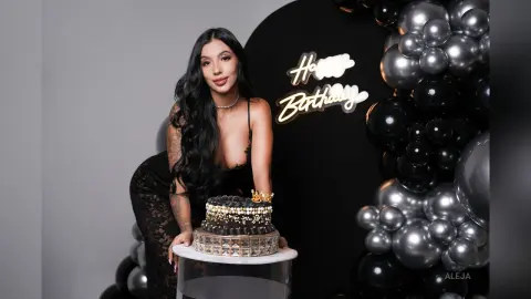 Aleja poses behind a black and gold birthday cake, wearing a black lace bodysuit with long wavy black hair.