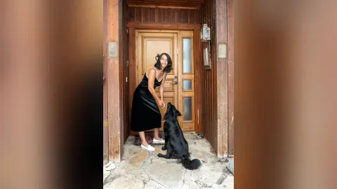 MichaelaBelle wears a black slip dress while standing in a wooden doorway next to a black dog.