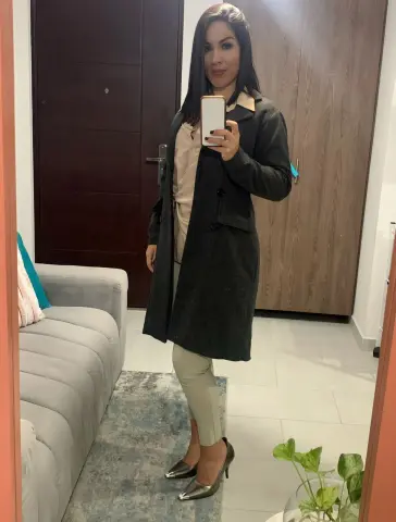 Woman in a long charcoal coat and beige trousers taking a mirror selfie in a modern living room.