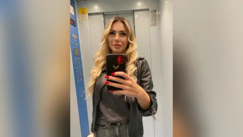 Blonde woman in a black leather jacket taking a selfie in an elevator mirror.