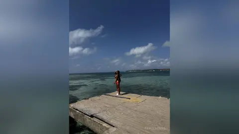 PamelaCreed stands on a wooden pier overlooking the ocean, wearing a red bikini or swimsuit with her hair down.