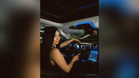 Young woman with long dark hair sitting in a luxury car interior with blue ambient lighting.