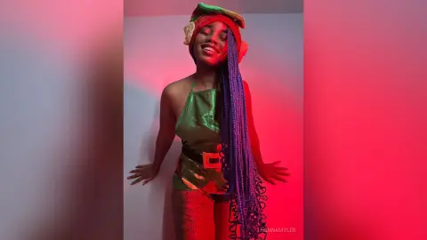Smiling woman in green superhero costume and long purple braids posing under vibrant red lighting.