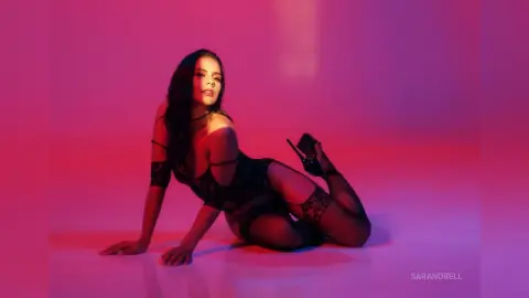 Glamorous woman in black lace lingerie sitting against a vibrant pink and red studio background.