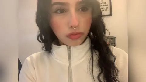 ZoeMonrow has long, wavy dark hair and wears a white zip-up turtleneck or jacket while looking directly at the camera.