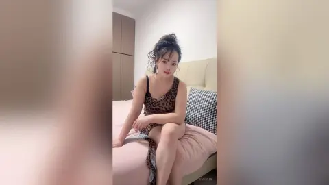 EnyaLin sits on a pink bed wearing a leopard print camisole dress with her hair in a messy bun.