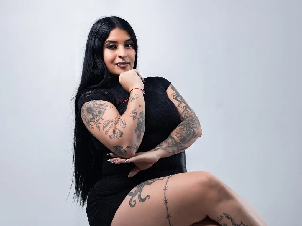 BellaMonrow poses with long black hair, wearing a black sleeveless dress that shows off her heavy arm and leg tattoos against a plain white background.