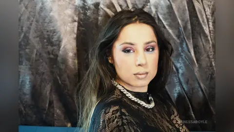 Woman with long dark hair and glamorous makeup wearing a black lace top against a textured silver backdrop.