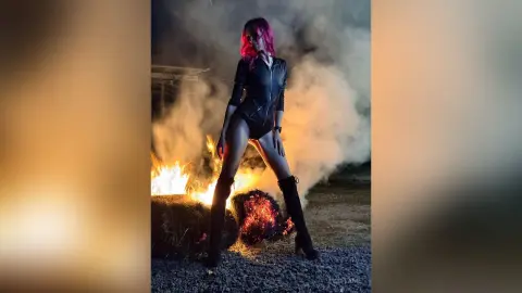 Pink-haired woman in a black bodysuit standing amidst dramatic smoke and fire in a dark outdoor setting.